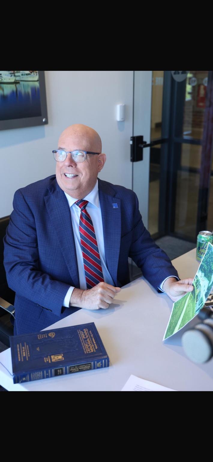 A seated interview guest holding a magazine at a table with books and a microphone